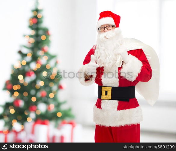 christmas, holidays, gesture and people concept - man in costume of santa claus with bag showing thumbs up over living room with tree