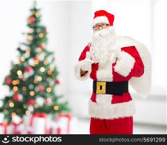 christmas, holidays, gesture and people concept - man in costume of santa claus with bag showing thumbs up over living room with tree