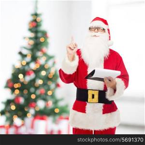 christmas, holidays, gesture and people concept - man in costume of santa claus with notepad pointing finger up over living room with tree background