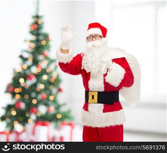 christmas, holidays, gesture and people concept - man in costume of santa claus with bag waving hand over living room with tree