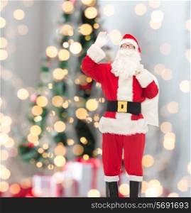 christmas, holidays, gesture and people concept - man in costume of santa claus with bag waving hand over tree lights background