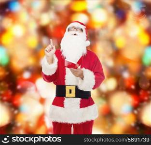 christmas, holidays, gesture and people concept - man in costume of santa claus with notepad pointing finger up over red lights background