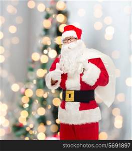 christmas, holidays, gesture and people concept - man in costume of santa claus with bag showing thumbs up over tree lights background