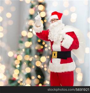 christmas, holidays, gesture and people concept - man in costume of santa claus with bag pointing finger up over tree lights background