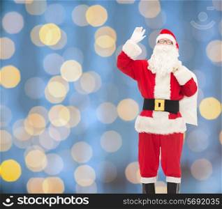 christmas, holidays, gesture and people concept - man in costume of santa claus with bag waving hand over blue lights background