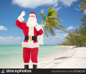 christmas, holidays, gesture and people concept - man in costume of santa claus with bag waving hand over tropical beach background