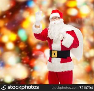 christmas, holidays, gesture and people concept - man in costume of santa claus with bag waving hand over red lights background
