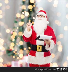 christmas, holidays, gesture and people concept - man in costume of santa claus with notepad pointing finger up over tee lights background