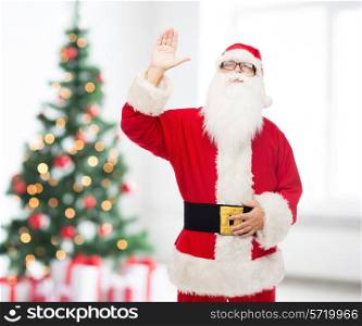 christmas, holidays, gesture and people concept - man in costume of santa claus waving hand over living room with tree background