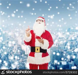 christmas, holidays, gesture and people concept - man in costume of santa claus with notepad pointing finger up over snowy city background