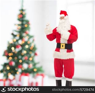 christmas, holidays, gesture and people concept - man in costume of santa claus pointing fingers over living room and tree background