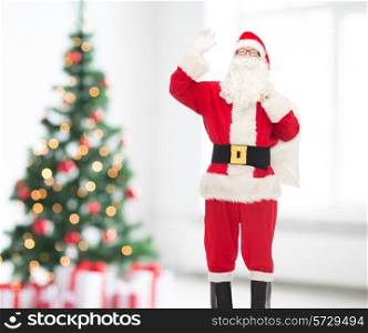 christmas, holidays, gesture and people concept - man in costume of santa claus with bag waving hand over living room and tree background
