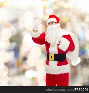 christmas, holidays, gesture and people concept - man in costume of santa claus with bag waving hand over lights background