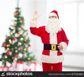 christmas, holidays, gesture and people concept - man in costume of santa claus waving hand over living room and tree background