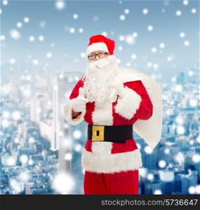 christmas, holidays, gesture and people concept - man in costume of santa claus with bag showing thumbs up over snowy city background