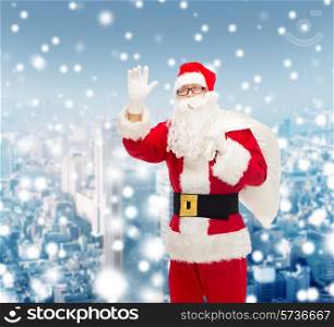 christmas, holidays, gesture and people concept - man in costume of santa claus with bag waving hand over snowy city background