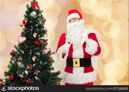 christmas, holidays, gesture and people concept - man in costume of santa claus with bag and christmas tree showing thumbs up over beige lights background