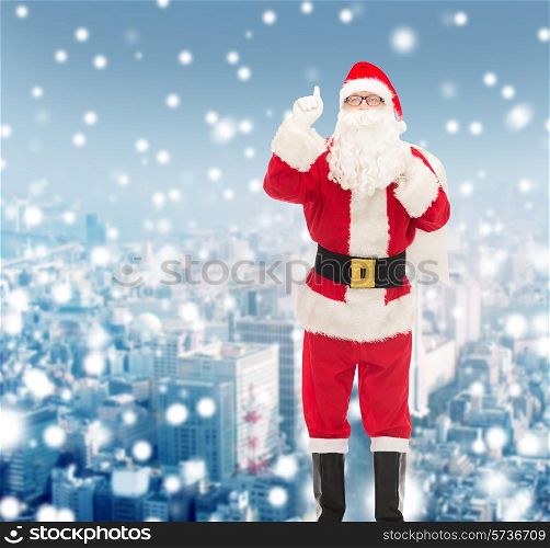 christmas, holidays, gesture and people concept - man in costume of santa claus with bag pointing finger up over snowy night city background