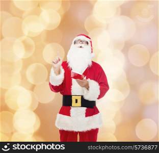 christmas, holidays, gesture and people concept - man in costume of santa claus with notepad pointing finger up over beige lights background