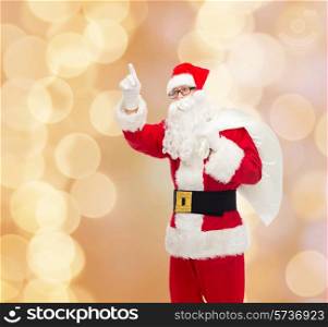 christmas, holidays, gesture and people concept - man in costume of santa claus with bag pointing finger up over beige lights background