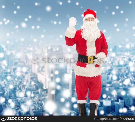 christmas, holidays, gesture and people concept - man in costume of santa claus waving hand over snowy city background