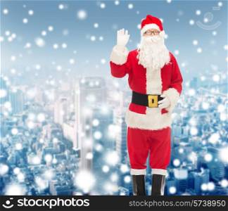 christmas, holidays, gesture and people concept - man in costume of santa claus waving hand over snowy city background