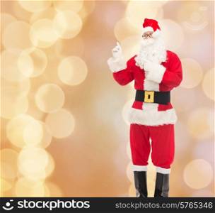 christmas, holidays, gesture and people concept - man in costume of santa claus pointing fingers over beige lights background