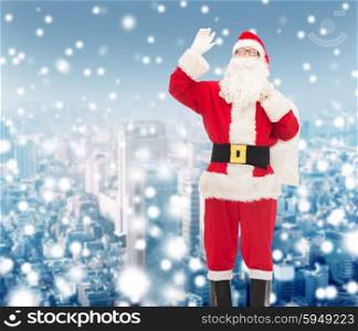 christmas, holidays, gesture and people concept - man in costume of santa claus with bag waving hand over snowy city background