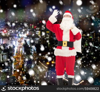 christmas, holidays, gesture and people concept - man in costume of santa claus with bag waving hand over snowy night city background