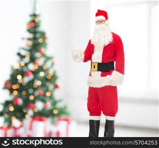 christmas, holidays, gesture and people concept- man in costume of santa claus showing thumbs up over living room and tree background