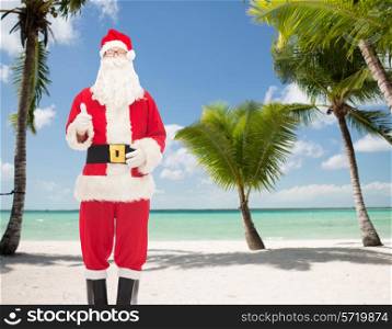 christmas, holidays, gesture and people concept- man in costume of santa claus showing thumbs up over tropical beach background