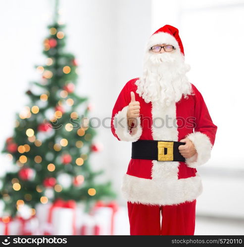 christmas, holidays, gesture and people concept- man in costume of santa claus showing thumbs up over living room with tree background