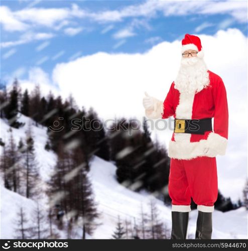christmas, holidays, gesture and people concept- man in costume of santa claus showing thumbs up over snowy mountains background