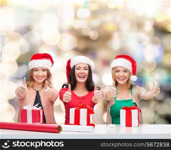 christmas, holidays, gesture, decoration and people concept - smiling women in santa helper hats with decorating paper and gift boxes showing thumbs up over lights background