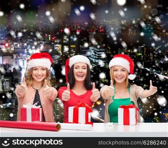 christmas, holidays, gesture, decoration and people concept - smiling women in santa helper hats with decorating paper and gift boxes showing thumbs up over snowy night city background