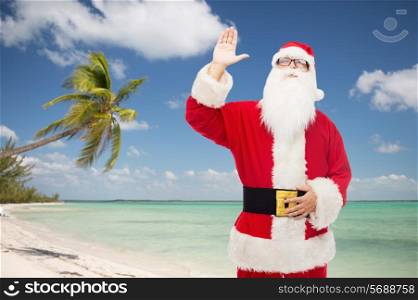 christmas, holidays, gesture, travel and people concept - man in costume of santa claus waving hand over tropical beach background
