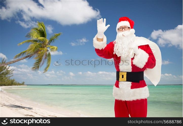 christmas, holidays, gesture, travel and people concept - man in costume of santa claus with bag waving hand over tropical beach background