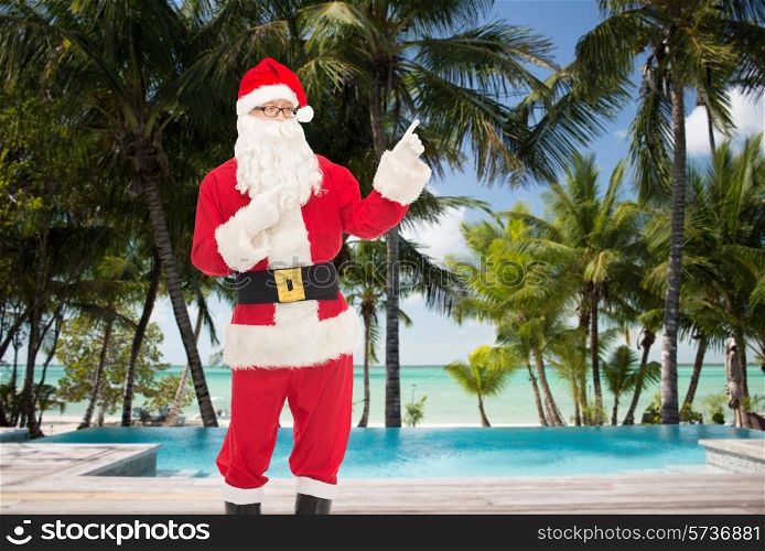 christmas, holidays, gesture, travel and people concept - man in costume of santa claus pointing fingers over swimming pool on tropical beach background