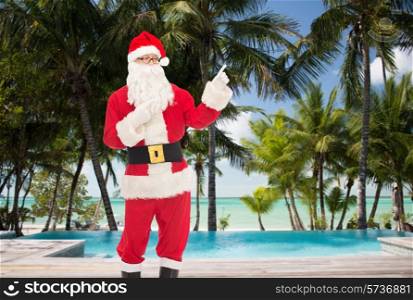christmas, holidays, gesture, travel and people concept - man in costume of santa claus pointing fingers over swimming pool on tropical beach background