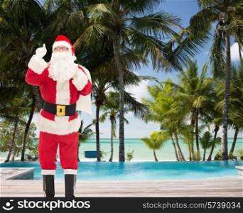 christmas, holidays, gesture, travel and people concept - man in costume of santa claus with bag pointing finger up over tropical beach and swimming pool background