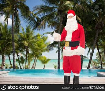 christmas, holidays, gesture, travel and people concept- man in costume of santa claus showing thumbs up over tropical beach and swimming pool background