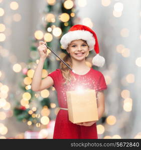 christmas, holidays, happiness and people concept - smiling girl in santa helper hat with gift box and magic wand over living room and christmas tree background