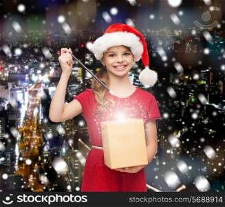 christmas, holidays, happiness and people concept - smiling girl in santa helper hat with gift box and magic wand over snowy night city background