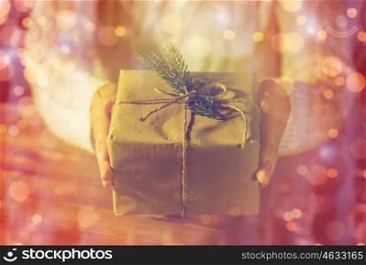 christmas, holidays, presents, new year and people concept - close up of woman hands holding gift box or parcel wrapped into brown mail paper and decorated with fir brunch over lights