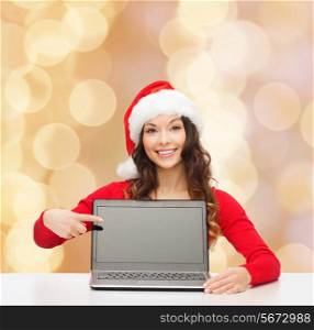 christmas, holidays, technology, advertisement and people concept - smiling woman in santa helper hat pointig finger to blank laptop computer screen over beige lights background