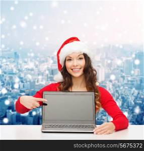 christmas, holidays, technology, advertisement and people concept - smiling woman in santa helper hat pointig finger to blank laptop computer screen over snowy city background