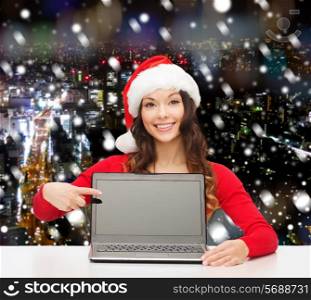 christmas, holidays, technology, advertisement and people concept - smiling woman in santa helper hat pointig finger to blank laptop computer screen over snowy night city background
