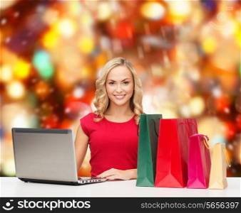 christmas, holidays, technology, advertising and people concept - smiling woman in red blank shirt with shopping bags and laptop computer over red lights background