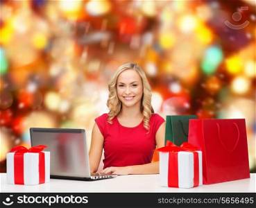 christmas holidays, technology, advertising and people concept - smiling woman in red blank shirt with shopping bags, gifts and laptop computer over red lights background