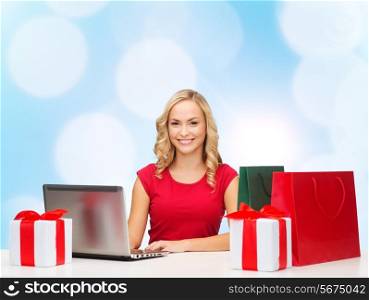 christmas, holidays, technology, advertising and people concept - smiling woman in red blank shirt with shopping bags, gifts and laptop computer over blue lights background